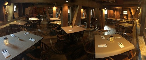 Cozy restaurant interior with wooden beams, tables set with utensils, and warm lighting.