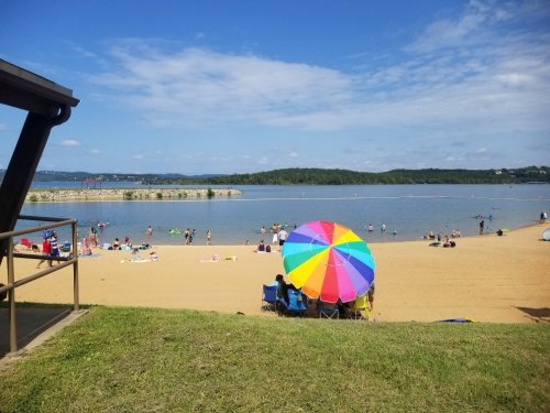 A sunny beach scene with people swimming and relaxing, featuring a colorful umbrella on the sand by the water.