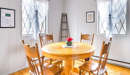 A cozy dining area with a round wooden table, four chairs, and a vase of flowers, illuminated by natural light.