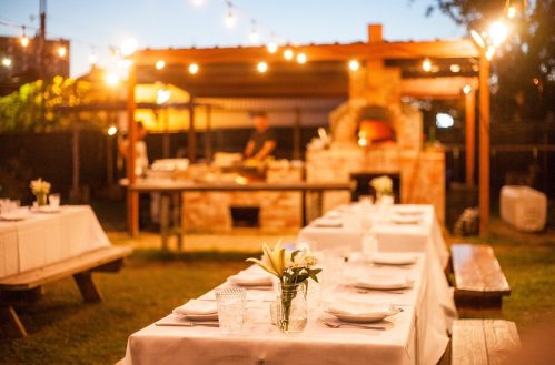 Outdoor dining setup with tables, flowers, and string lights, featuring a brick oven in the background at dusk.