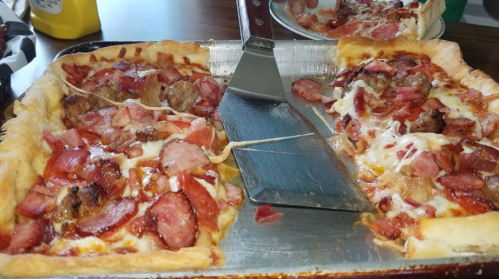 A close-up of a pizza with a thick crust, topped with cheese, bacon, and sausage, with a slice being lifted.