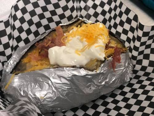 A baked potato topped with cheese, bacon bits, and sour cream, served in a checkered paper container.