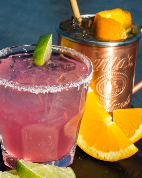 Two colorful cocktails: a pink drink with a lime wedge and a copper mug filled with a dark beverage and orange slices.