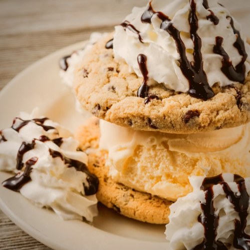 A dessert featuring two chocolate chip cookies sandwiching vanilla ice cream, topped with whipped cream and chocolate drizzle.
