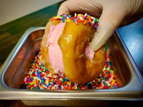 A hand in a glove holds a donut filled with pink frosting, surrounded by colorful sprinkles in a metal tray.