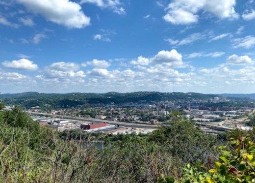 A panoramic view of a cityscape with green hills, blue skies, and fluffy clouds in the background.