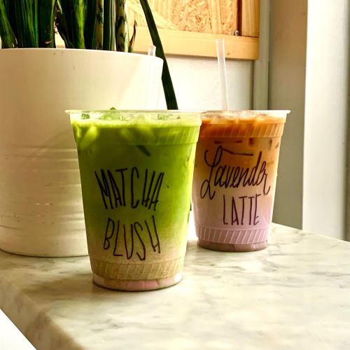 Two colorful drinks on a marble surface: a green matcha blush and a lavender latte, both in clear cups with straws.