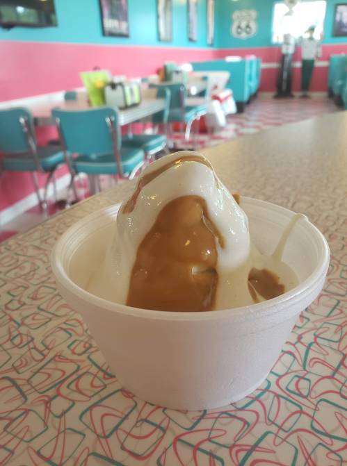 A cup of ice cream topped with caramel sauce, set on a retro diner table with colorful decor in the background.