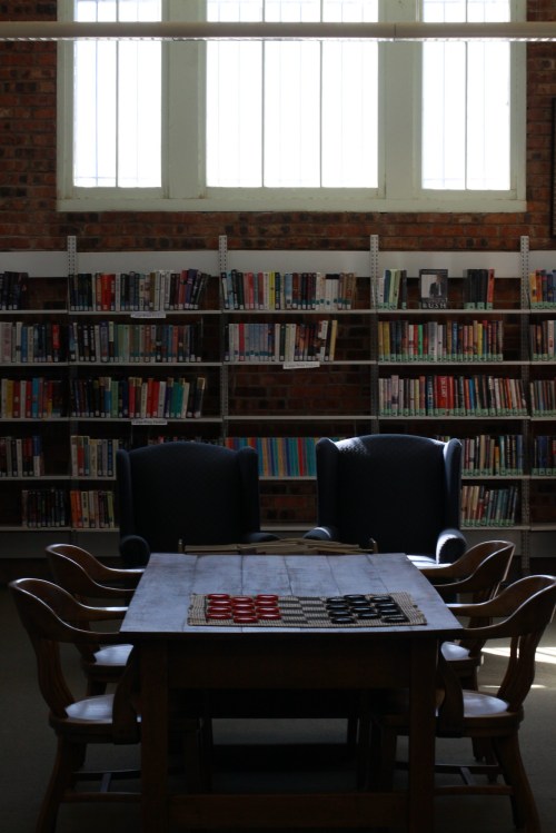 A cozy library scene featuring a wooden table with checkers, two chairs, and bookshelves filled with books.