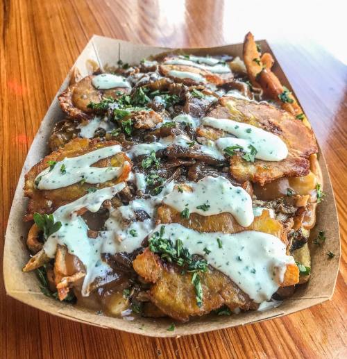 A wooden tray filled with crispy plantains topped with sauce and herbs, served on a wooden table.
