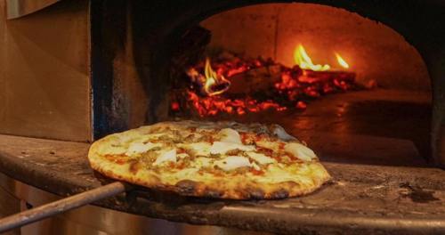 A freshly baked pizza being pulled from a wood-fired oven, with flames and glowing embers in the background.
