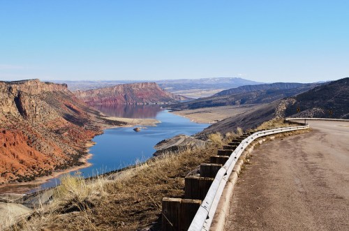 Scenic view of a winding road alongside a river, surrounded by red rock formations and distant mountains under a clear blue sky.