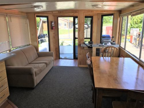 Interior of a cozy cabin with a sofa, dining table, and large windows overlooking a deck and outdoor area.