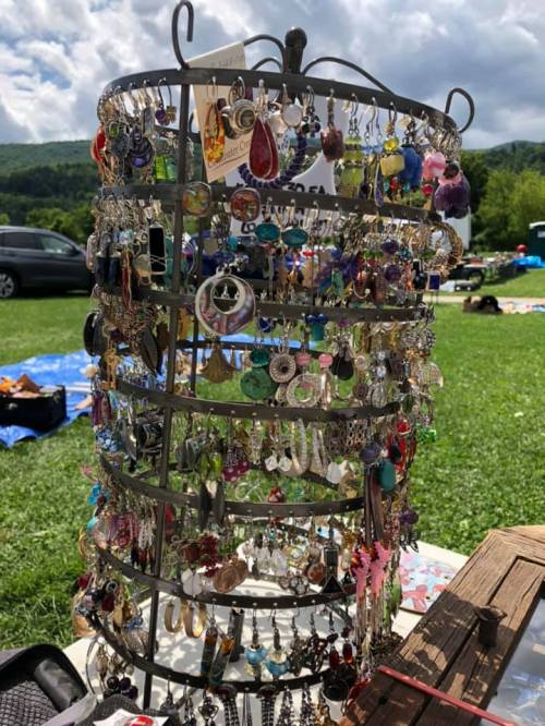 A rotating display rack filled with colorful earrings and jewelry, set outdoors with a green landscape in the background.