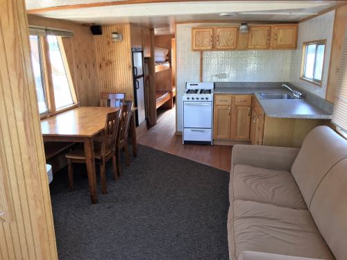 Interior of a cozy cabin featuring a dining table, sofa, kitchen with stove, and a view of a bedroom area.