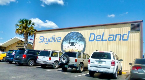 A building with "Skydive DeLand" painted on the side, surrounded by parked cars and a palm tree under a blue sky.