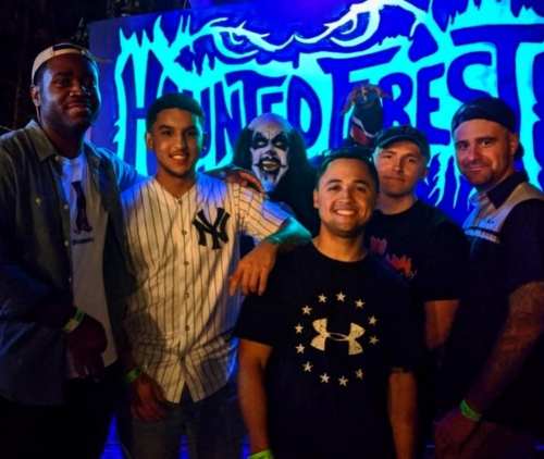 A group of six friends poses together in front of a colorful haunted forest backdrop, smiling and enjoying the moment.