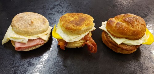 Three breakfast sandwiches: an English muffin with ham and egg, a biscuit with bacon and egg, and a croissant with egg and cheese.