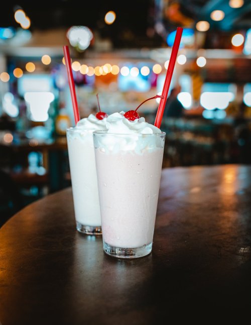 Two milkshakes in tall glasses, one pink and one white, topped with whipped cream and cherries, on a wooden table.