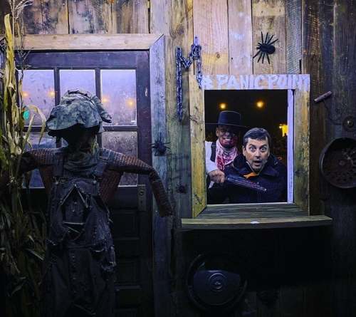 A surprised man looks out from a spooky window, with a scarecrow and creepy decorations nearby.