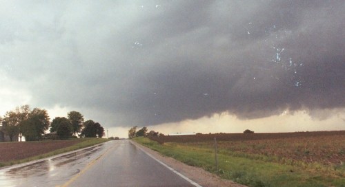 A dark, stormy sky looms over a wet rural road lined with fields and trees.