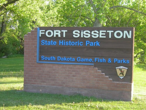 Sign for Fort Sisseton State Historic Park, featuring blue text on a wooden background, surrounded by greenery.