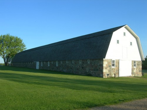 A long, white barn with a stone base, set against a clear blue sky and green grass.