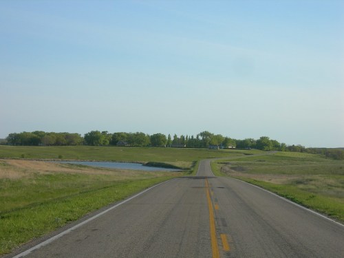 A winding road leads through green fields, with a pond visible on the left and trees in the background under a clear sky.