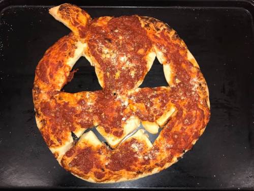 A pizza shaped like a jack-o'-lantern with a smiling face and a stem, baked to a golden brown.