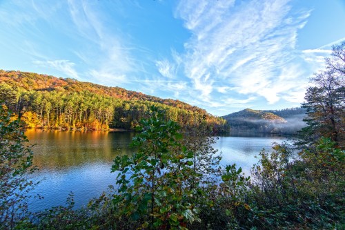 A serene lake surrounded by colorful autumn trees under a blue sky with wispy clouds.