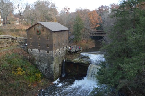 A historic wooden mill beside a waterfall, surrounded by trees with autumn foliage and people on a viewing platform.
