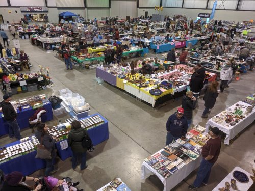 A bustling indoor market with various vendor tables, shoppers browsing, and colorful displays of goods.