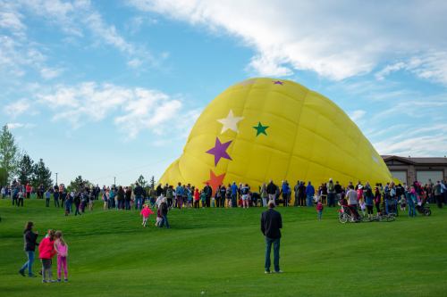 A large yellow hot air balloon with colorful stars is being prepared, surrounded by a crowd on a grassy field.