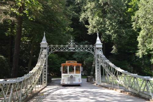 A vintage trolley crosses a decorative bridge surrounded by lush green trees.