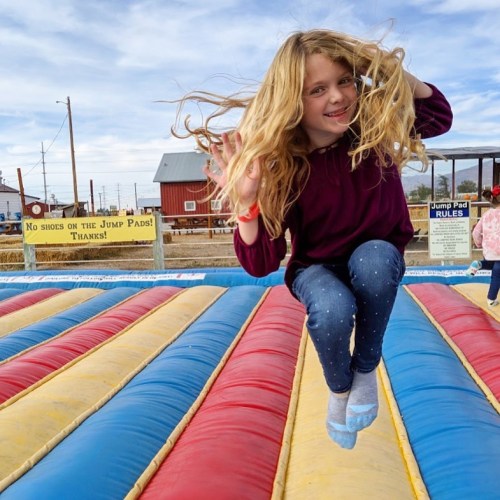 A girl with long hair jumps joyfully on a colorful inflatable bounce pad outdoors.