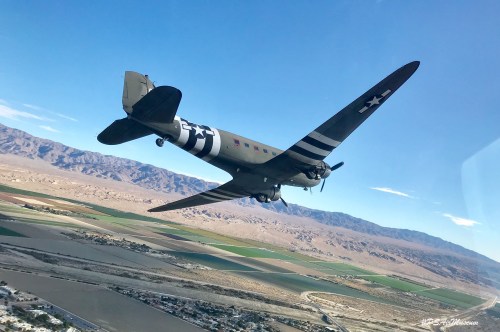 A vintage aircraft flying over a mountainous landscape and fields, showcasing its distinctive military paint scheme.