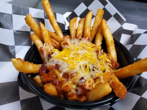 A bowl of loaded fries topped with chili and melted cheese, served in a black container with a checkered paper lining.