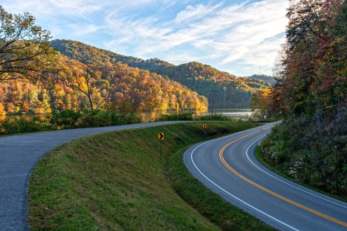 A winding road curves alongside a lake, surrounded by vibrant autumn foliage and rolling hills under a blue sky.