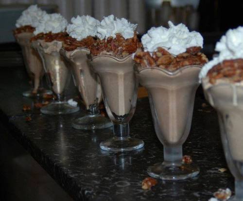 Six dessert cups filled with chocolate mousse, topped with whipped cream and crushed nuts, arranged on a dark countertop.