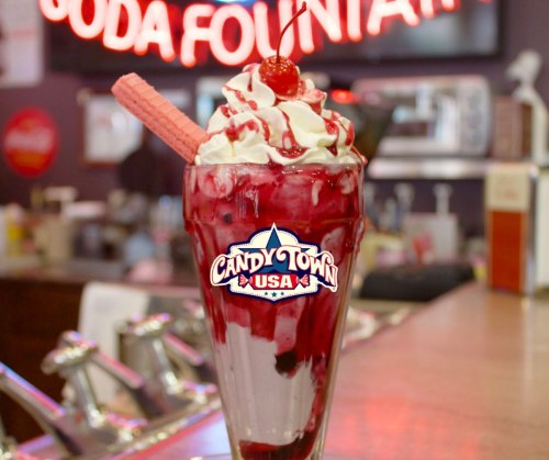 A tall sundae glass filled with ice cream, cherry topping, whipped cream, and a wafer, with a neon sign in the background.
