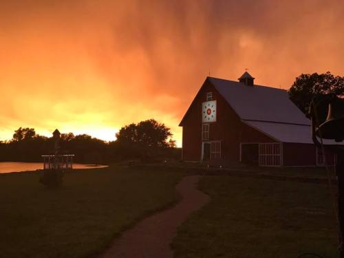 A red barn with a clock on its side stands against a vibrant orange sunset, with a path leading to a nearby pond.