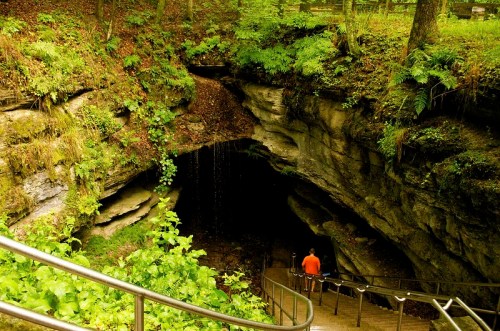 A person in an orange shirt descends stairs into a dark cave surrounded by lush greenery and rock formations.
