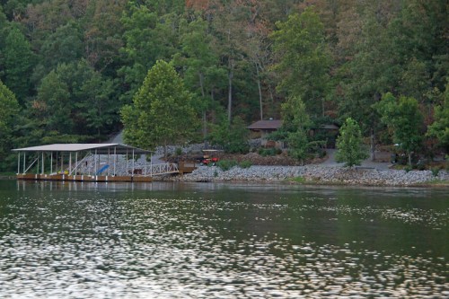A serene lakeside view with a dock, trees, and a cabin surrounded by lush greenery.