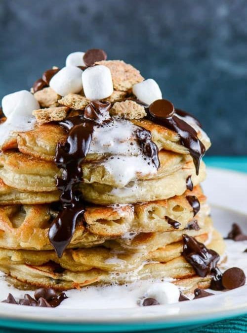 A tall stack of pancakes topped with chocolate sauce, marshmallows, and graham cracker pieces on a white plate.
