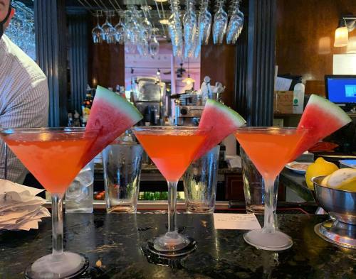 Three vibrant cocktails with watermelon slices on the rim, set against a bar backdrop with glassware.