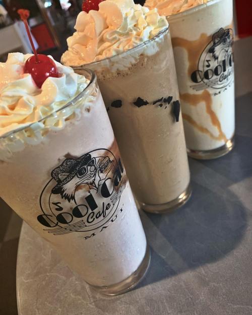 Three milkshakes in tall glasses, topped with whipped cream and cherries, displayed on a table.