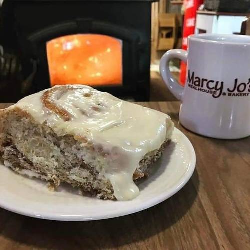 A slice of cinnamon roll with cream cheese frosting on a plate, next to a coffee mug, with a warm fireplace in the background.