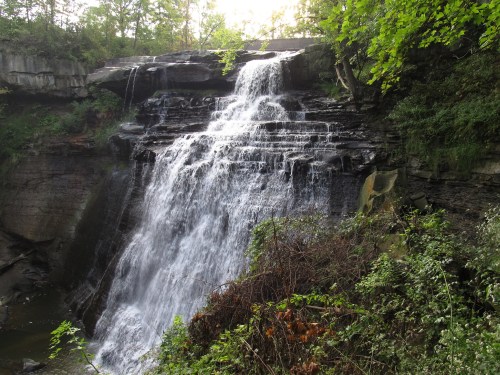 A cascading waterfall flows over rocky cliffs, surrounded by lush greenery and trees in a serene natural setting.
