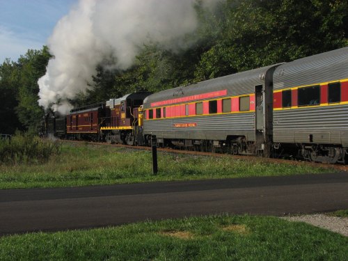 A vintage steam train billows smoke as it travels along a scenic route, with passenger cars in tow.