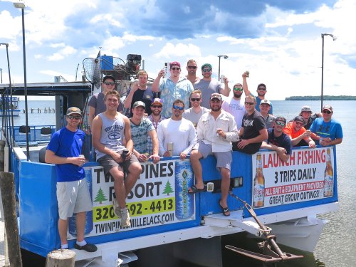 A large group of people poses on a fishing boat, smiling and holding drinks, with a scenic waterfront in the background.